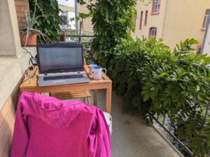 Laptop auf einem Tisch auf dem Balkon meines Schreibzimmers in Trossingen, daneben eine Kletterpflanze am Haus – hier schreibe und redigiere ich als Autorin.
